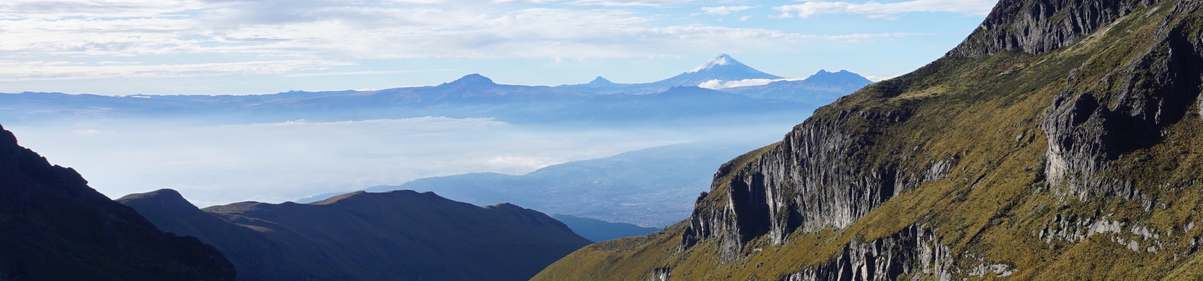 Una fotografía corta y ancha mirando hacia el horizonte desde un punto de vista elevado en las laderas del volcán Pichincha. En la distancia, se ve el cono nevado del volcán Antisana, y en primer plano hay acantilados cubiertos de hierba.