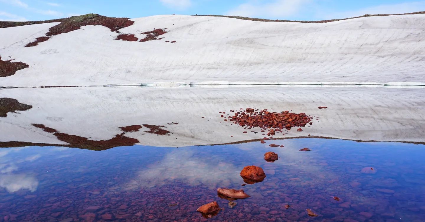 A photograph of the caldera lake of Mount Azhdahak, with the snowy rim reflected in still water with stepping stones.