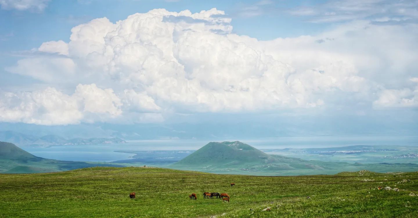 Huge cumulonimbus clouds form over Lake Sevan, viewed a distance across the grassy slopes of the Gegham Range over some horses and a volcanic cone.