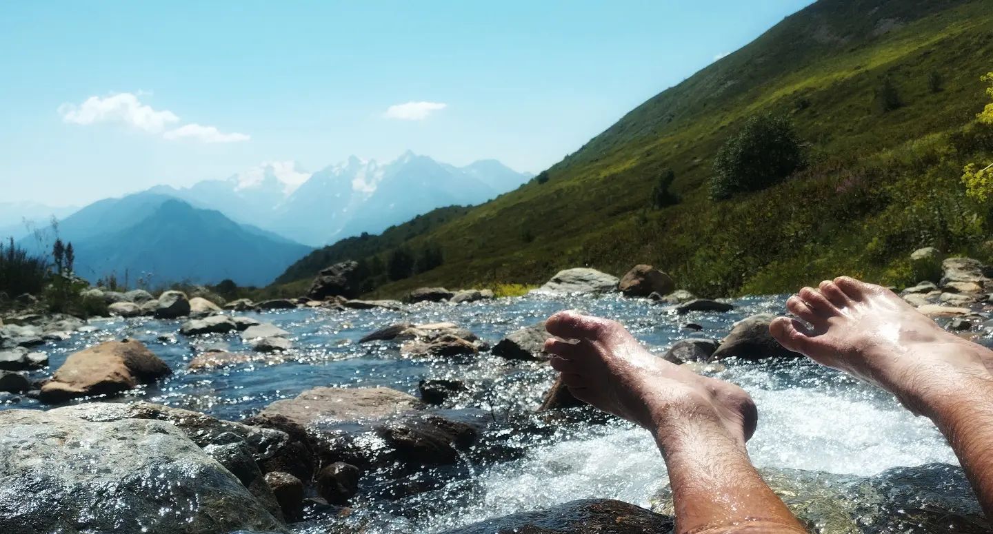 A photograph taken in a stream close to the water level, looking at the hiker’s feet with mountains in the background.