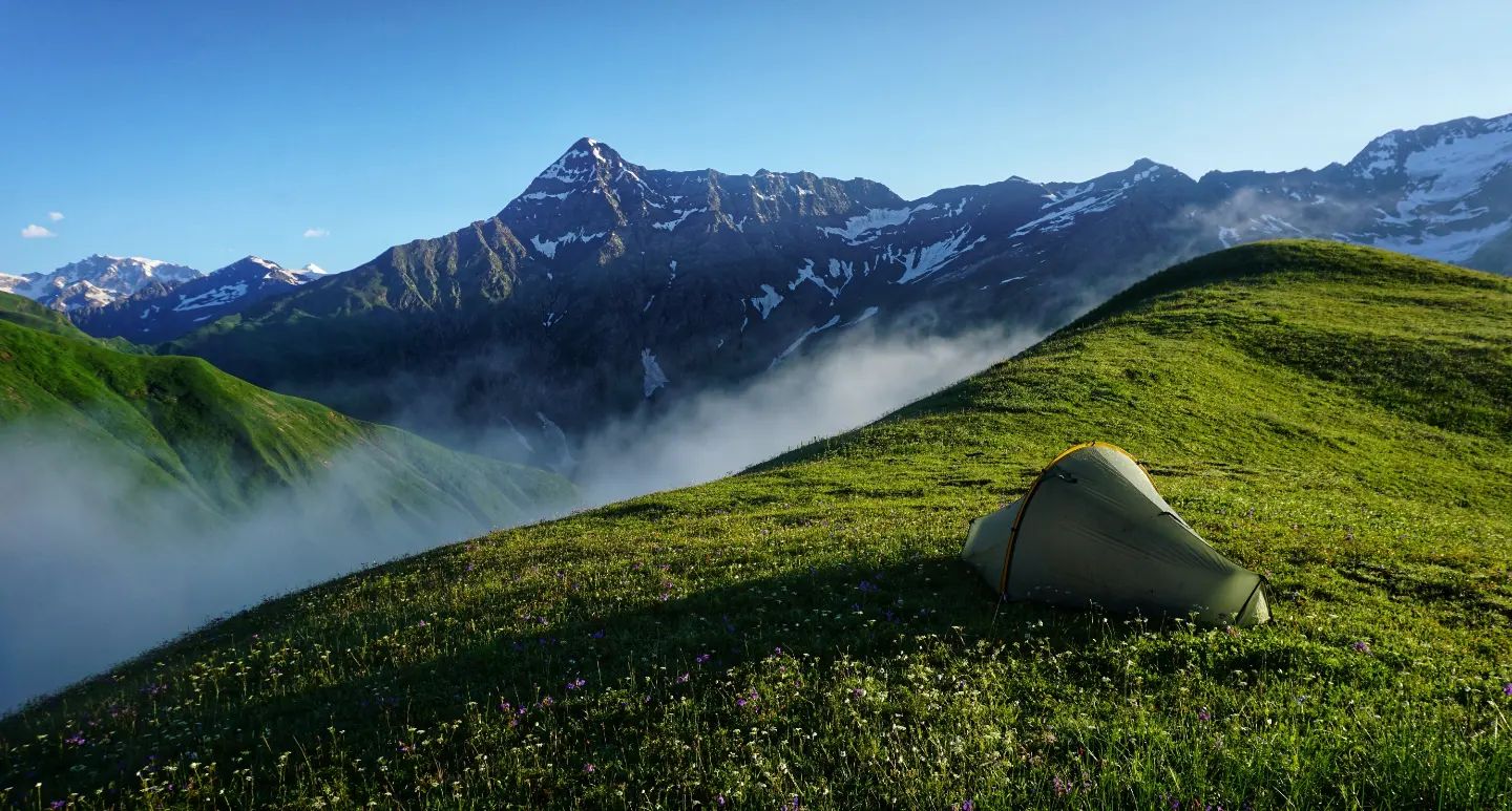 A one-person tent pitched on the grassy Sasvano Ridge, with the snowy main ridge of the Greater Caucasus in the background.