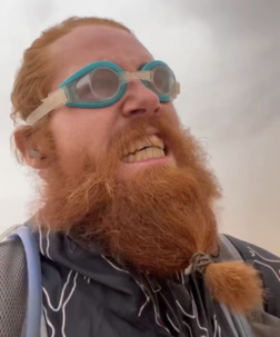 An image of Russ Cook taken from one of his Instagram videos filmed during day 337 of his journey. The shot is of Cook’s face and bushy ginger beard. Cook is grimacing and wearing swimming goggles as he is running through a Saharan dust storm.