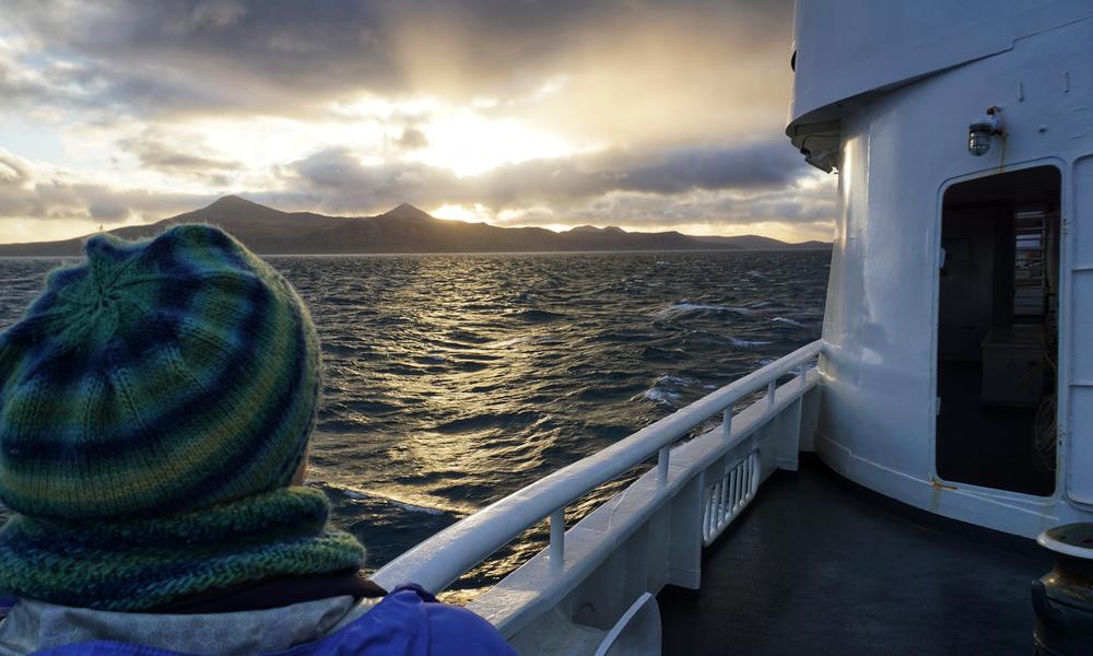A photograph of the ocean, taken from the deck of a large metal boat. The sun is setting behind an island ridge, and reflecting on a choppy sea. The sky is partially cloudy. In the foreground, the back of a person’s head is visible and they are wearing a colourful beanie hat. On the right of the image, the boat’s deck, railing, and a doorway are all visible.