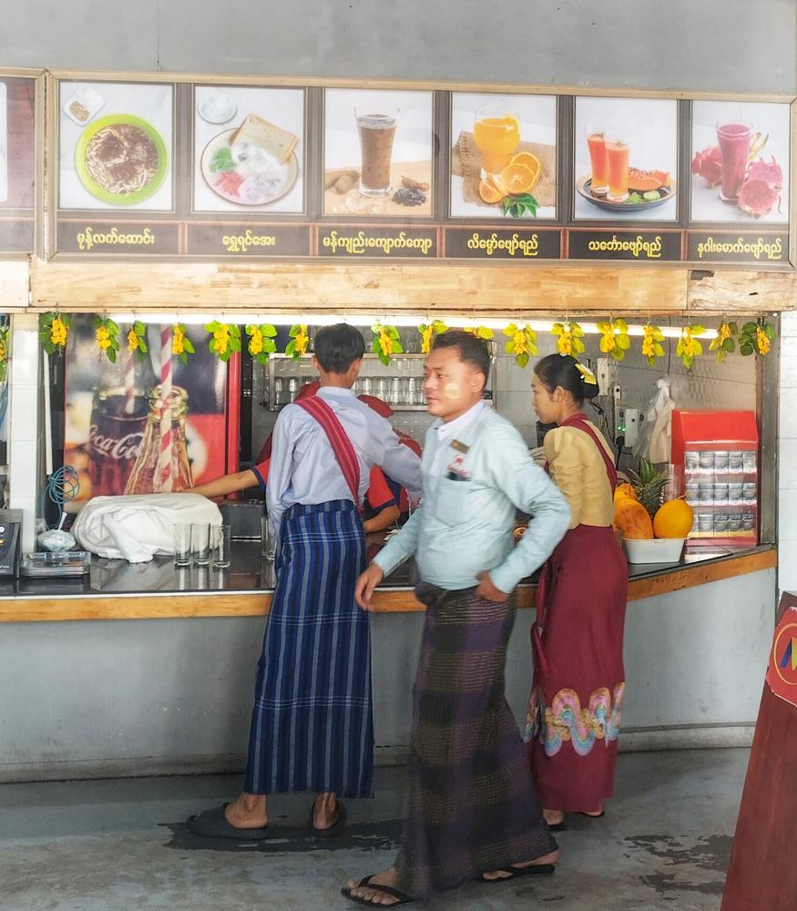 A photo of counter serving refreshments in Myanmar. Two men and one woman are visible in front of the counter, and all are wearing the traditional longyi (a garmet like a dress, consisting of a single piece of fabric wrapped around the waist and extending to the ankles) and sandals. The face of one of the men is visible and on his cheeks he has of thanaka, a paste made from a root, worn as skincare. Above the counter are pictures of juices and snacks labelled in Burmese.