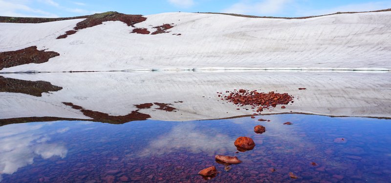Una fotografía del lago de la caldera del Monte Azhdahak, con el borde nevado reflejado en agua quieta con piedras de paso.