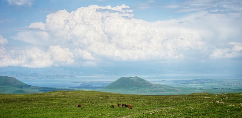 Huge cumulonimbus clouds form over Lake Sevan, viewed a distance across the grassy slopes of the Gegham Range over some horses and a volcanic cone.
