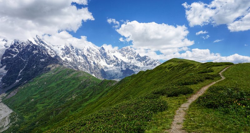 Una fotografía de la pared nevada del macizo de Shkhara, vista desde el mirador de Chkhunderi, que es una cresta verde con un sendero bien pisado.
