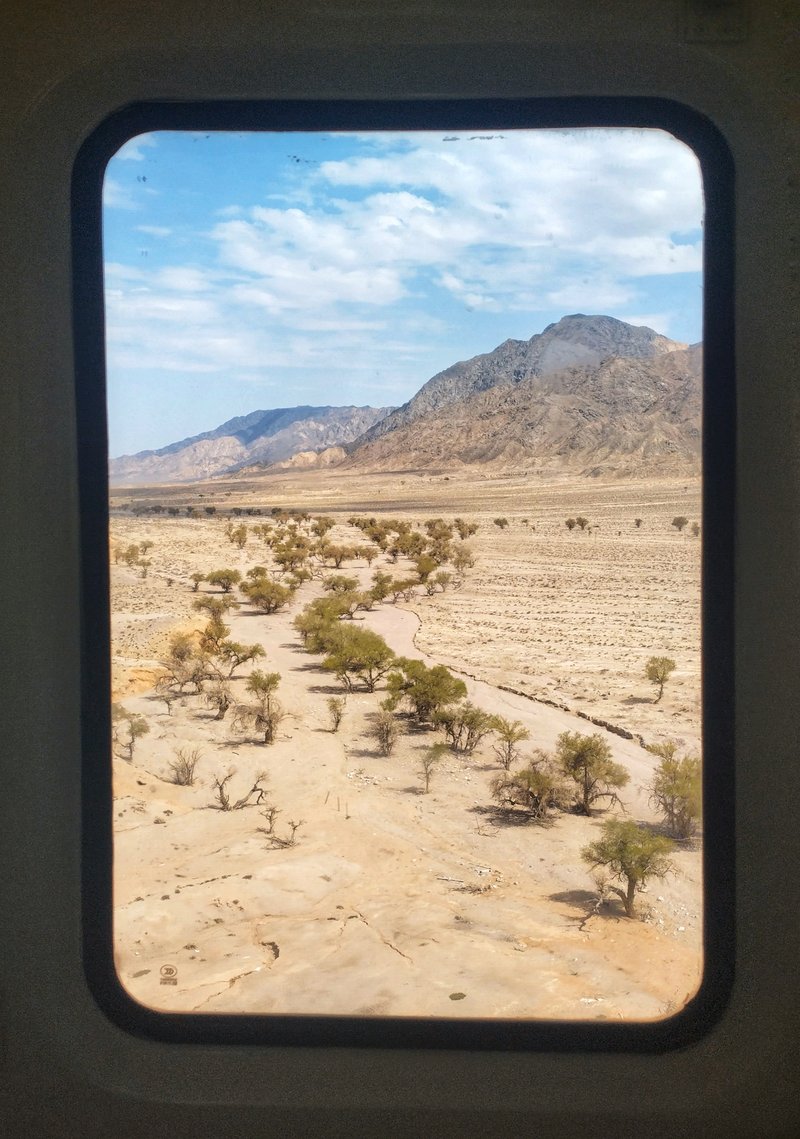 A photograph framed by the window of a train, showing a dry river with some trees and a mountainous, arid landscape.