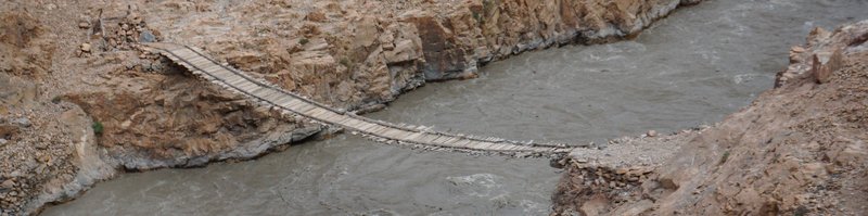 A wide-format photograph of a hand-made footbridge over the Bartang River, Tajikistan.